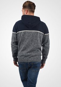 Sweat à capuche en marine foncé et gris, avec un tricot texturé. Présente une bande horizontale contrastante aux épaules et des bords-côtes.