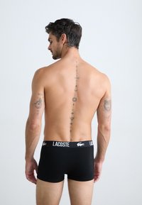 Boxer shorts noirs avec un logo Lacoste blanc sur la ceinture. Le modèle a des tatouages visibles le long de la colonne vertébrale et du bras.