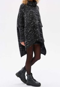 Schwarz-weißer melierter Strick-Poncho mit hohem Kragen, oversized Design und seitlichen Schlitzen; getragen über schwarzen Shorts und Strumpfhosen, kombiniert mit klobigen schwarzen Stiefeln.