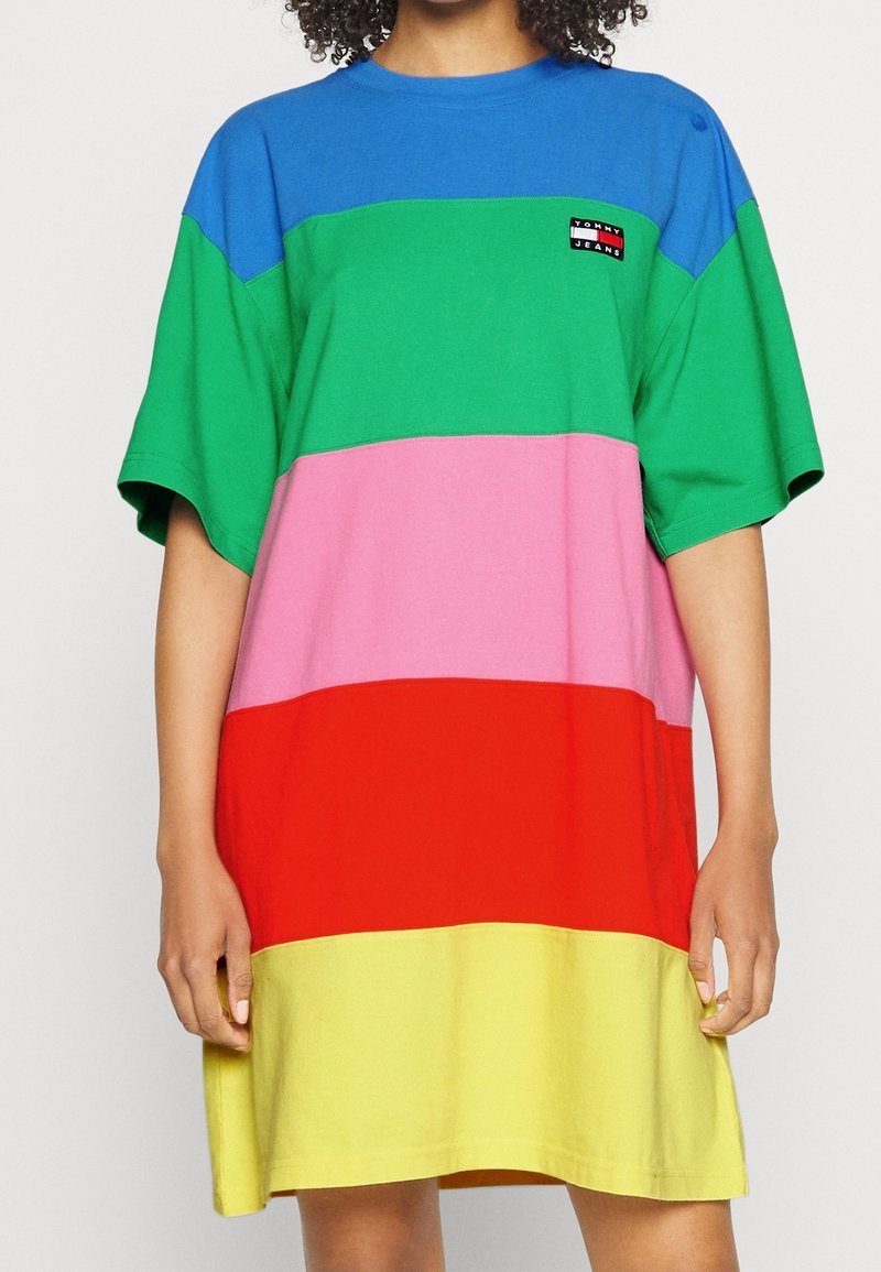 Personne portant une robe ample à manches courtes avec des blocs de couleurs horizontaux en bleu, vert, rose, rouge et jaune, avec un petit patch logo.