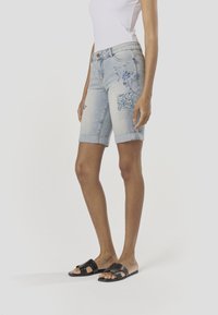 Világos koptatású farmer rövidnadrág virágos hímzéssel és kék mintás folttal. Felhajtott szegélyek és testhez simuló szabás. Fekete papucs.