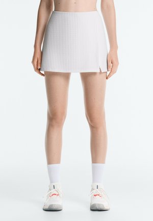 Person, die einen weißen, strukturierten Minirock mit seitlichem Schlitz, weiße Knöchelsocken und weiße Sportschuhe trägt und vor einem einfarbigen Hintergrund steht.