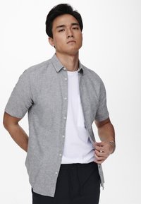 Camisa de botón de manga corta en un gris claro, hecha de tejido tejido, sobre una camiseta blanca. Presenta un cuello clásico y un detalle de bolsillo.
