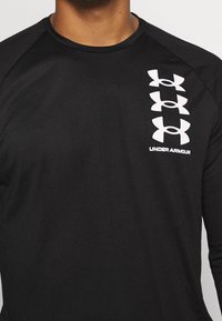 Czarna koszulka z długim rękawem, wykonana z gładkiego materiału. Posiada wiele białych logotypów pionowo ułożonych po lewej stronie. Okrągły dekolt.