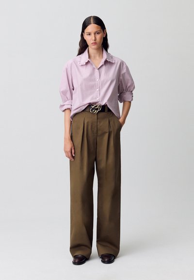 Camisa de botones a rayas rosas con mangas oversized y cuello, combinada con pantalones marrones de cintura alta y un cinturón negro decorativo.