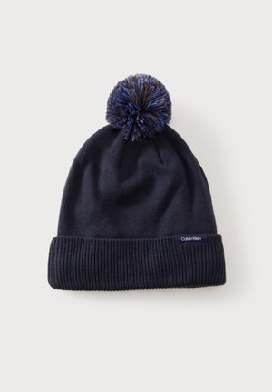Marineblaue gestrickte Wintermütze mit geripptem Bündchen und mehrfarbigem Bommel oben, mit kleinem Calvin Klein Etikett am Bündchen.