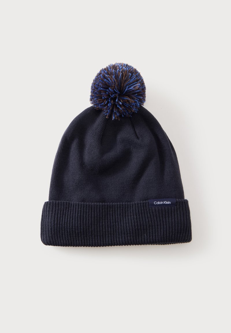 Bonnet d'hiver en maille bleu marine avec bord-côte et pompon multicolore sur le dessus, avec une petite étiquette Calvin Klein sur le bord.