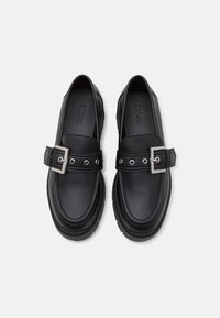 Paire de mocassins en cuir noir avec boucles argentées et détails de couture, estampillés "HUGO" sur les semelles intérieures, vus de dessus.