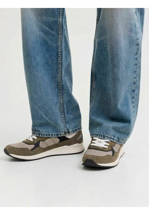 Personne portant un jean bleu clair et des baskets vertes et beiges avec des lacets blancs, debout sur une surface claire et uniforme.