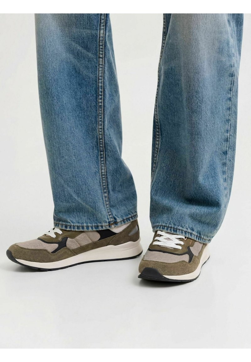 Personne portant un jean bleu clair et des baskets vertes et beiges avec des lacets blancs, debout sur une surface claire et uniforme.