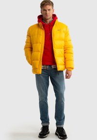 Geltona pūkinė striukė su siuvinėtu paviršiumi, dėvima virš raudonos džemperio, derinama su mėlynomis džinsais ir juodais žygio batais. Modelis stovi stovintis veidu.