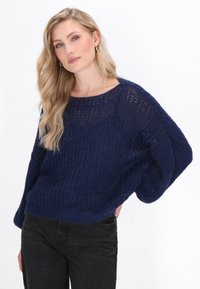 Marineblauer Strickpullover mit lockerer Passform, gerippter Textur und weiten Ärmeln. Kombiniert mit schwarzen Jeans. Der Hintergrund ist schlicht weiß.