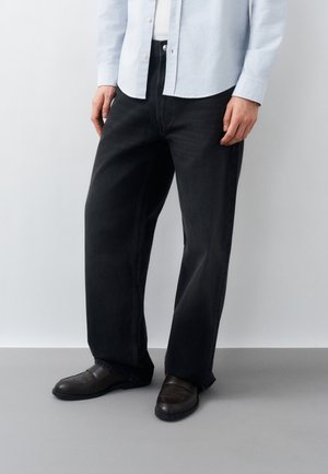 Mężczyzna ubrany w czarne szerokie jeansy, czarne skórzane lordsy, białą koszulę i jasnoniebieską koszulę zapinaną na guziki, stojący na szarej podłodze.