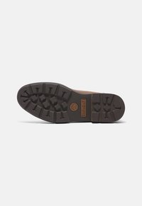 Ruskea kumipohja, jossa on syviä uramalleja ja Timberland-logo. Ominaisuuksiin kuuluu pyöreä muoto ja kestävä rakenne, jotka tarjoavat pitoa ja kestävyytä.