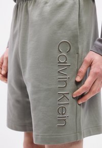 Κοντινό πλάνο ανοιχτόγκρι σορτς Calvin Klein με ελαστική μέση και κάθετο λογότυπο στο αριστερό πόδι, φορεμένα από άτομο με το χέρι κοντά στην τσέπη.