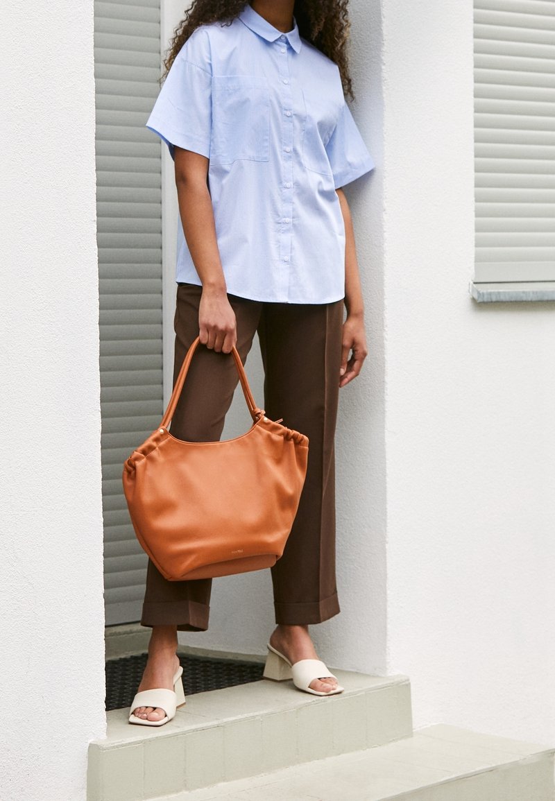 Borsa a tracolla in pelle marrone con design arricciato, color arancione, e manici corti, abbinata a una camicia button-up azzurro chiaro e pantaloni marroni.