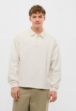 Młody mężczyzna w kremowej, długorękawowej koszulce polo i beżowych spodniach, stojący na tle białego, gładkiego tła.