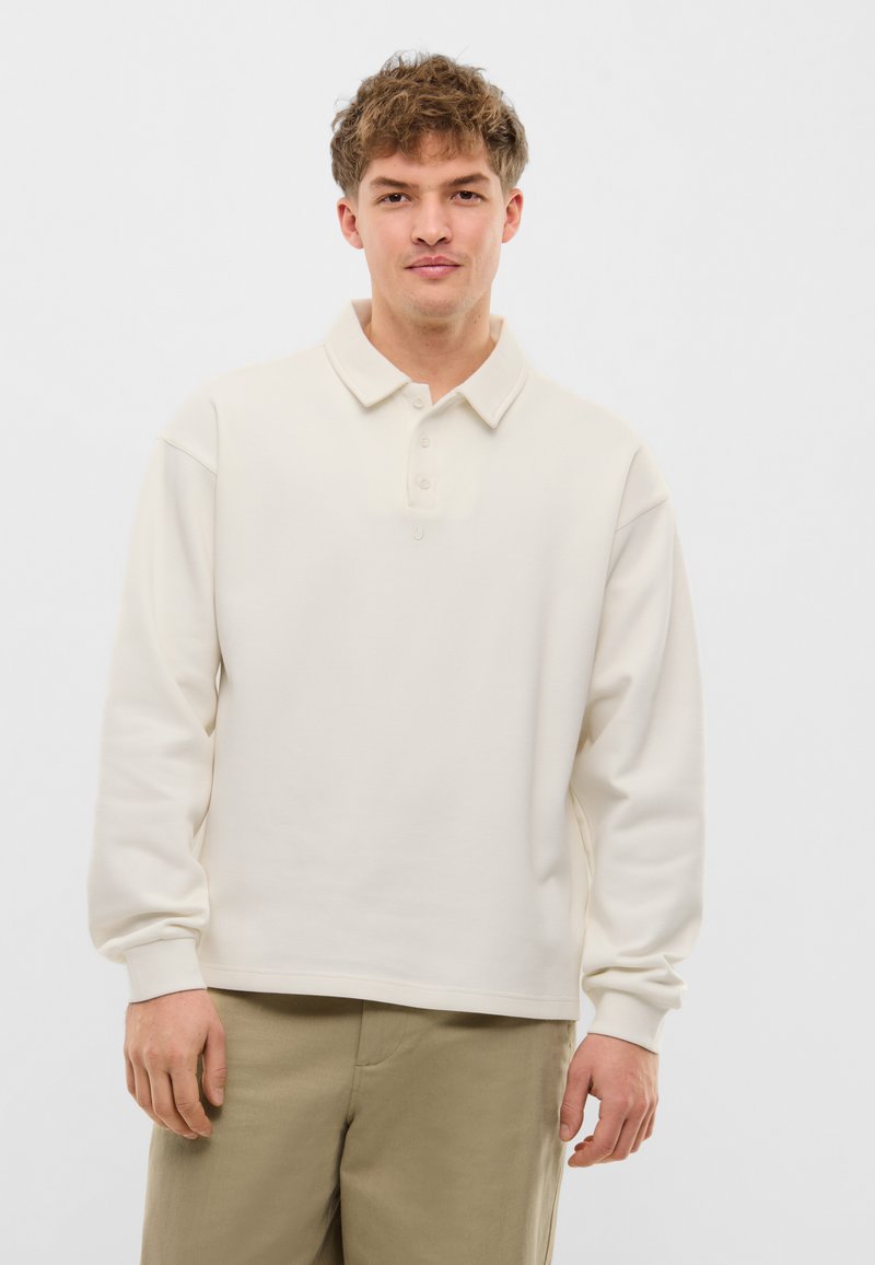 Ung mand iført en cremefarvet poloshirt med lange ærmer og beige bukser, stående mod en ensartet hvid baggrund.