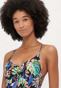 Black swimsuit with multicolored tropical leaf pattern, adjustable straps, and a ruched detail at the center. Soft, smooth fabric texture.