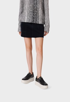 Persona che indossa una minigonna nera, un maglione argentato con paillettes e sneakers piattaforma nere con suola bianca spessa, in piedi davanti a uno sfondo neutro.