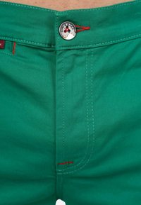Pantaloni turchesi con chiusura zip, dotati di un bottone argentato con il marchio "Chpo&Baux" e accenti di cucitura rossa lungo le cuciture.