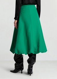 Grüner A-Linien-Rock aus glattem Stoff, mit mittellangem Saum und einem klaren Taillenbund, kombiniert mit hochglänzenden schwarzen kniehohen Stiefeln.
