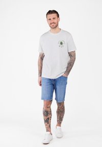 Hombre con tatuajes de manga completa y en las piernas, vestido con una camiseta blanca con un gráfico de palmera, pantalones cortos de mezclilla y zapatillas blancas, sonriendo con la mano en el bolsillo.