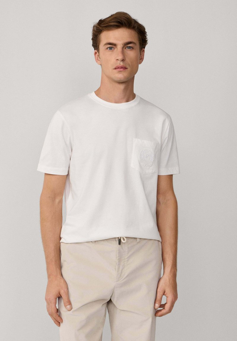 Camiseta blanca de algodón con cuello redondo y mangas cortas, que presenta un bolsillo texturizado con un diseño en relieve.