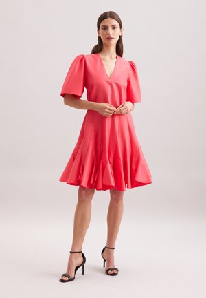 Robe corail avec des manches courtes bouffantes et un décolleté en V, dotée d'un ourlet évasé. Assortie à des sandales à talons hauts noires avec des lanières. Texture lisse.