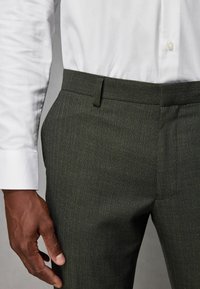 Mørkegrønne skræddersyede bukser med en subtil tekstur; flad front, slim fit og bæltestropper, parret med en hvid skjorte med knapper.