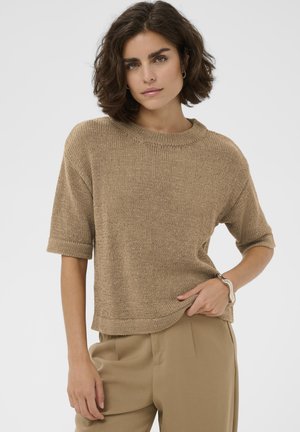 Donna con capelli corti e scuri che indossa un maglione a maniche corte color ruggine lavorato a maglia e pantaloni beige, posa con una mano in tasca su uno sfondo bianco.