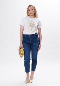 Hvid bomulds T-shirt med grafisk design og teksten "Girl Queen", parret med blå denimjeans, holdende en skinnende guldsmaakker og guld hæle.