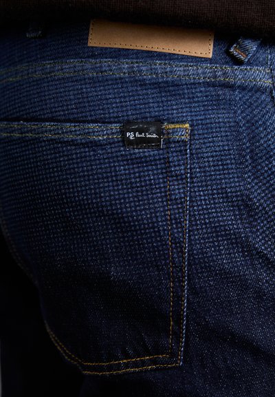 Jeans de denim azul oscuro con un acabado texturizado, con costuras amarillas y una etiqueta negra que dice "PS Paul Smith" en el bolsillo trasero.