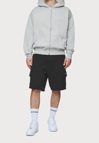 Felpa grigia con zip e tasche frontali, abbinata a pantaloni cargo neri con tasche laterali. Sneakers bianche e calzini sportivi con logo.