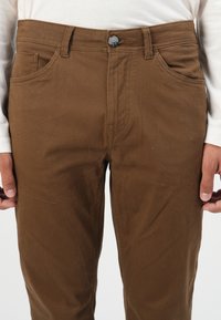 Man in een bruin broek en een wit langemouwen shirt, handen ontspannen langs de zijden, close-up op de taille en bovenbenen.