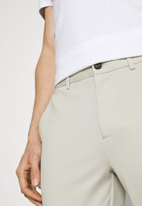 Gros plan d'une personne portant un pantalon beige clair texturé avec un bouton noir et une chemise blanche à manches courtes, main détendue sur le côté.