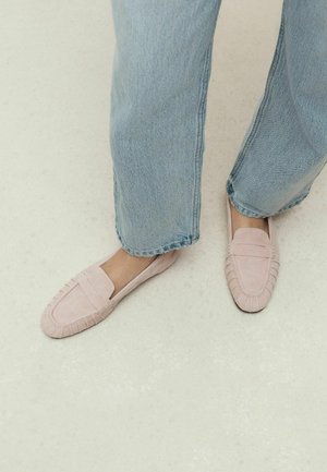 Person trägt hellblaue Jeans und hellrosa Wildleder-Loafer und steht auf einem hellen Boden.