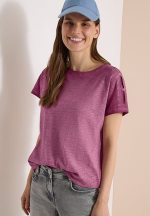 Lachende vrouw met een lichtblauwe pet, paarse top met korte mouwen en schouderdetail, en een grijze spijkerbroek tegen een neutrale achtergrond.