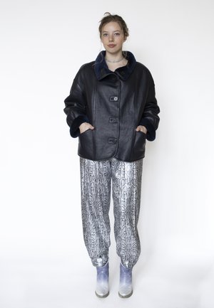 Jeune personne les mains dans les poches, vêtue d'une veste en cuir noire avec bordure en fourrure, d'un pantalon imprimé serpent et de bottines à gradient argenté vers bleu.