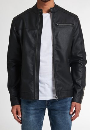 Mann trägt schwarze Lederjacke mit Reißverschluss über weißem T-Shirt und blauen Jeans, steht mit entspannten Armen an der Seite.