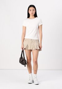 Hvit kortermet t-skjorte, beige volang mini skjørt, hvite høye sneakers, og en liten mørk veske i hånden mot en ensfarget bakgrunn.