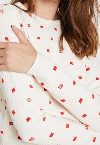 Weißer Texturopullover mit einem Rautenmuster und roten quadratischen Akzenten sowie einem Logo. Rippbündchen am Kragen und an den Ärmeln sind sichtbar.