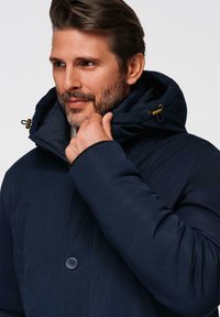 Marineblauwe geïsoleerde jas met een capuchon, voorzien van een ritssluiting aan de voorkant, knoopsluiting en textielstof. De capuchon heeft verstelbare trekkoorden.