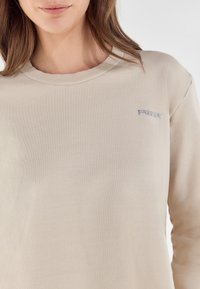 Felpa beige a maniche lunghe con collo a costine e discreto logo "Freddy" in glitter argento sulla zona sinistra del petto. Tessuto morbido e design semplice.