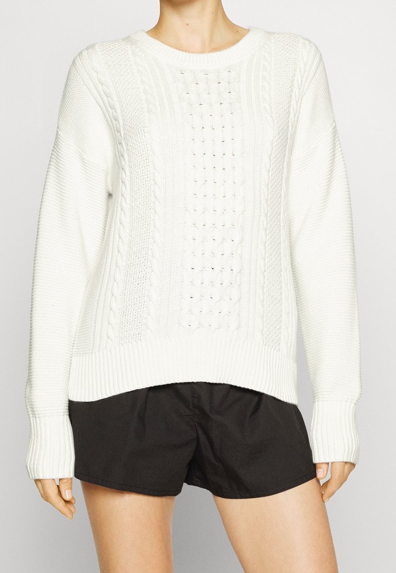 Kremowy sweter z wzorem warkocza, okrągłym dekoltem, ściągaczami na rękawach i na dole; noszony na czarnych szortach, prezentujący fakturowaną tkaninę.