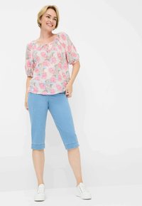 Blusa floreale con motivi rosa, blu e beige, vestibilità comoda, scollo a cuore, abbinata a pantaloni capri azzurri e sneakers bianche.
