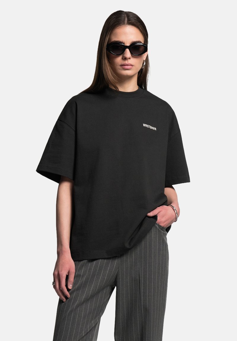 Černé oversized tričko s kulatým výstřihem a krátkými rukávy, se bílým textovým logem. V kombinaci se šedými kalhotami s jemným proužkem.