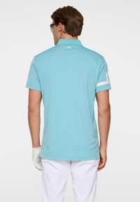Homme portant un polo bleu clair avec un logo blanc sur le col et la manche, un pantalon blanc, un gant blanc, vu de dos sur un fond uni.