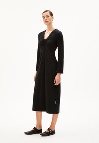 Schwarzes langärmliges Kleid mit V-Ausschnitt und Frontbindedetail, mit lockerer Passform und knielanger Länge, kombiniert mit schwarzen Ballerinas.