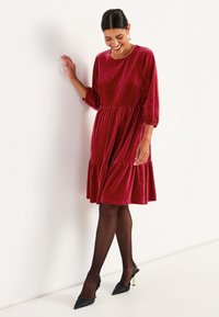 Rotes Samtkleid mit Dreiviertelärmeln und gestuftem Rock. Getragen mit schwarzen transparenten Strumpfhosen und spitzen Absätzen. Glatte Textur und runder Ausschnitt.